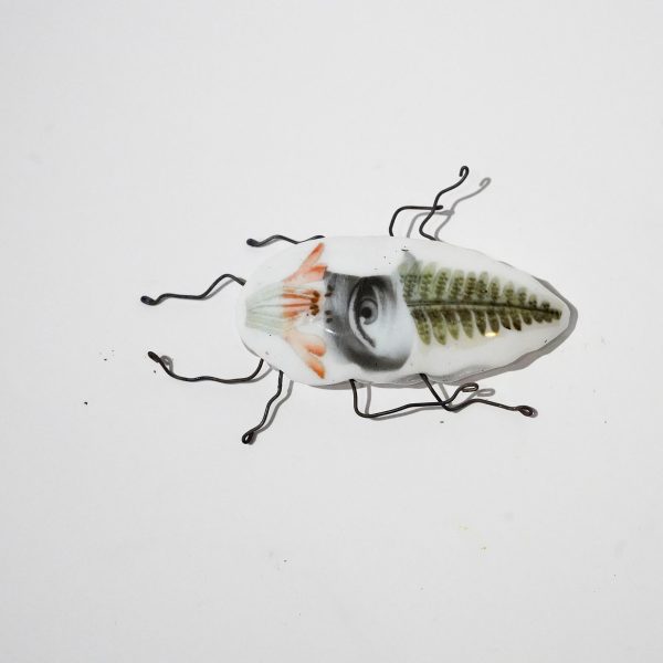 Biały szklany chrząszcz zdobiony zdjęciem oka i paproci. Chrząszcz ma nogi z drutu.