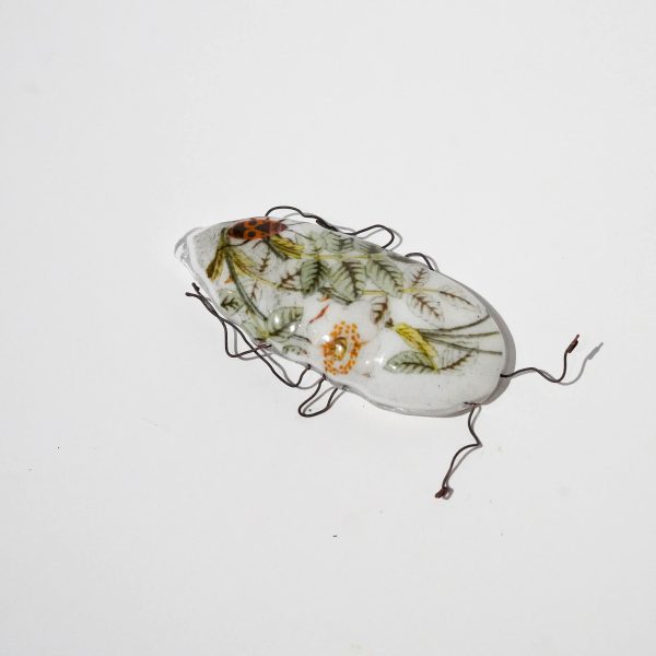 Biały szklany chrząszcz zdobiony grafiką z listkami i czerwonym robakiem. Chrząszcz ma nogi z drutu.