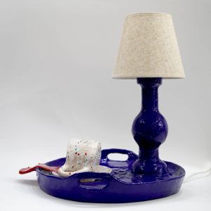 Lampa z postawą w kolorze ultramaryny, z abażurem z tkaniny w kolorze kremowym. Podstawa lampy jest w kształcie tacy z przewróconą białą filiżanką.